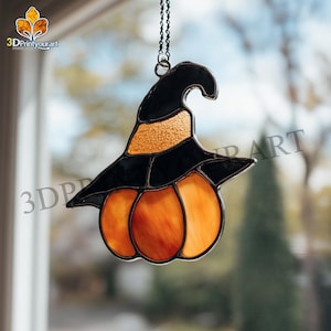Könnte beinhalten: Halloween-Dekoration aus Glas mit einem Hexenhut auf einem Kürbis. Der Hut ist schwarz mit einem goldfarbenen, strukturierten Band, und der Kürbis ist orange. Die Dekoration hängt an einer Kette vor einem verschwommenen Hintergrund.