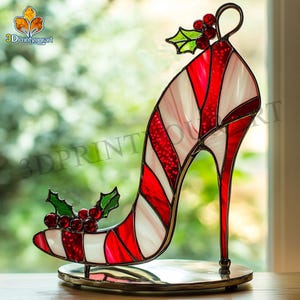 Peut inclure: Une chaussure à talon haut en verre à rayures rouge et blanche en forme de canne à sucre avec un accent de houx et de baies sur le dessus. La chaussure est sur un support argenté.