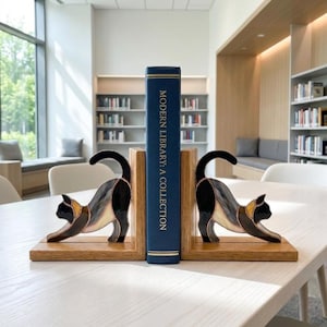 May include: A pair of wooden bookends featuring stained glass cat silhouettes in black, white, and amber tones, positioned on either side of a blue book titled Modern Library: A Collection, with the cats in a stretching pose.