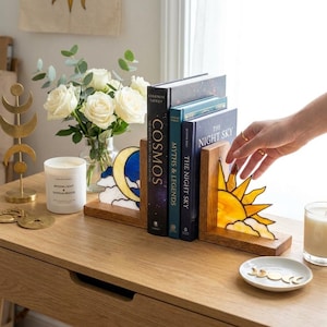 Op de afbeelding: Een paar houten boekensteunen met glas-in-lood inzetstukken, één met een gele maansikkel met witte wolken en de andere met een gele zon met witte wolken, die drie boeken ondersteunen met de titels Cosmos, Myths & Legends en The Night Sky.