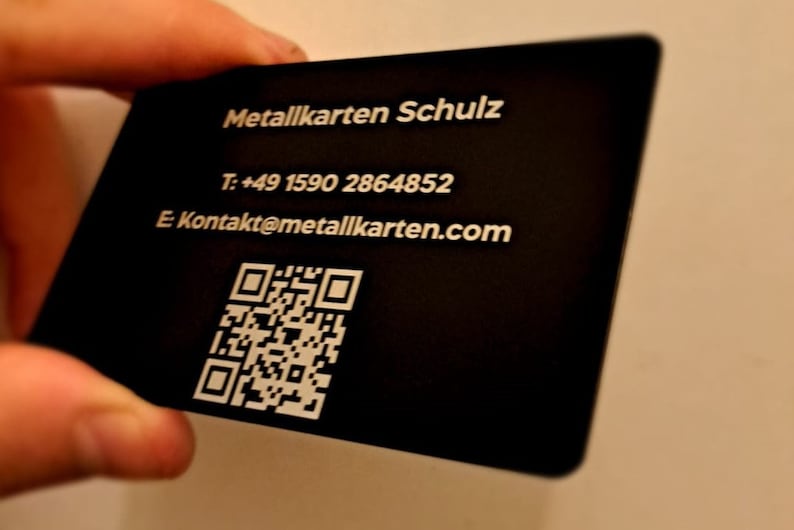 May include: A black metal business card with white text. The text reads 'Metallkarten Schulz', 'T: +49 1590 2864852', 'E Kontakt@metallkarten.com', and a QR code.