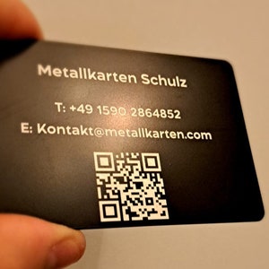 May include: A black metal business card with white text. The text reads 'Metallkarten Schulz', 'T: +49 1590 2864852', 'E: Kontakt@metallkarten.com' and a QR code.