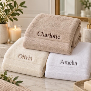 May include: Three folded cotton bath towels in beige, cream, and white. Each towel features a textured border and is embroidered with a name in a serif font: Charlotte on the beige towel, Olivia on the cream towel, and Amelia on the white towel.
