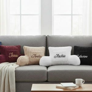 May include: A grey sofa with personalised corduroy backrest cushions. The cushions are burgundy, beige, white, and black, each embroidered with a name in cursive. Additional blue cushions and a cream throw blanket are also present.