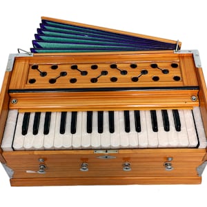May include: A wooden harmonium with a keyboard of black and white keys. The instrument has a light brown finish with decorative cutouts and a green and blue bellows. Metal corner protectors are visible.