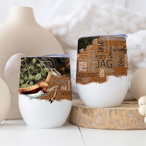 Puede incluir: Dos vasos de vino blancos sin pie. Uno presenta una ilustración de un springbok saltando con texto. El otro tiene un fondo marrón con texto. Ambos tienen una base blanca y un borde plateado.