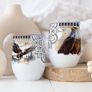 May include: Two white stemless wine tumblers with a watercolor design of an eagle. The design includes a black, tan, and white geometric pattern at the top. One tumbler shows an eagle in flight, and the other shows an eagle perched.