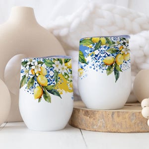 Op de afbeelding: Twee witte, stamloze wijnglazen met een citroen- en bloemenontwerp. Het ontwerp toont gele citroenen, witte bloemen en groene bladeren tegen een blauw-witte achtergrond. De glazen worden op een houten oppervlak gepresenteerd.