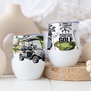 Puede incluir: Dos copas de vino blancas sin tallo con diseños temáticos de golf. Una presenta un carrito de golf, la otra tiene pelotas de golf, palos y el texto "Sorry if I looked interested, I was probably thinking about GOLF."