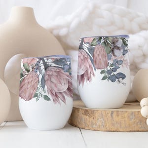 Könnte beinhalten: Zwei weiße Weingläser ohne Stiel mit floralen Designs. Die Gläser zeigen ein Aquarelldesign aus rosa Protea-Blüten, grünen Blättern und blauen Akzenten. Die Gläser stehen auf einer Holzoberfläche.