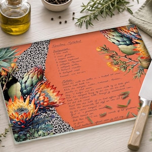 Puede incluir: Una tabla de cortar rectangular de vidrio con un centro naranja vibrante que presenta una receta impresa de Korstlose Melktert, rodeada de ilustraciones detalladas de flores de protea y un estampado de leopardo en blanco y negro.