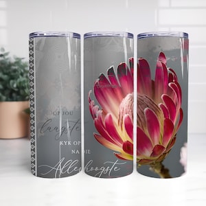 Puede incluir: Un vaso de acero inoxidable con un diseño de flor de protea en tonos rosa, rojo y amarillo sobre un fondo gris. El vaso también tiene texto en una fuente de escritura. El diseño envuelve todo el vaso.