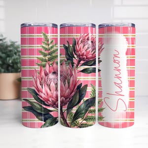 Può includere: Un bicchiere cilindrico con un motivo a quadri rosa e bianco e strisce dorate orizzontali. Il design presenta grandi fiori di protea rosa e foglie di felce verdi. Una pennellata bianca verticale contiene il nome Shannon in corsivo rosa.
