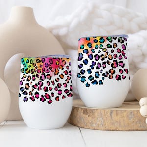 May include: Two white insulated wine tumblers with a colorful leopard print design. The print features a rainbow of colors, including pink, blue, yellow, and green. The tumblers are displayed on a wooden surface.