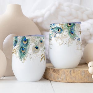 Può includere: Due bicchieri bianchi senza stelo con un motivo decorativo di piume di pavone nei toni del verde acqua, blu e oro. Il design avvolge la parte superiore di ogni bicchiere, mostrando piume dettagliate a forma di occhio e delicati accenti dorati.