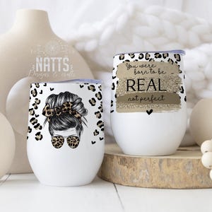 Puede incluir: Dos vasos de acero inoxidable blancos con un diseño de estampado de leopardo negro y dorado. Un vaso presenta a una mujer con un moño desordenado y gafas de sol. El otro vaso tiene el texto "You were born to be REAL not perfect".