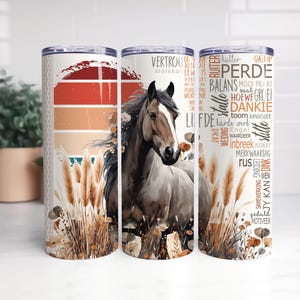 May include: A white tumbler with a brown horse and a background of sunset colors. The tumbler has a design of brown, orange, and white feathers. The tumbler has text in Dutch on the side.