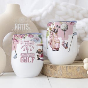 Op de afbeelding: Twee witte roestvrijstalen bekers met roze en bloemmotieven. De ene beker heeft een golfkarretje en de tekst "Sorry als ik geïnteresseerd leek, ik dacht waarschijnlijk aan GOLF". De andere beker heeft een roze golftas, golfclubs en een golfbal.
