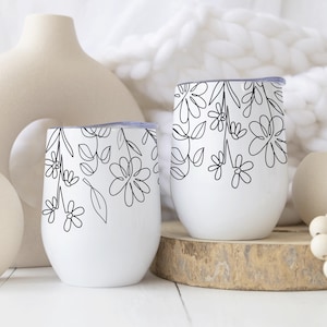 Puede incluir: Dos tazas de cerámica blanca con un dibujo de línea negra de flores y hojas. Las tazas están sobre una superficie de madera.