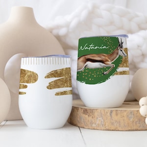 Op de afbeelding: Twee witte geïsoleerde wijnglazen. Eén heeft gouden abstracte vormen, de andere een groene achtergrond met een springende gazelle en de naam "Natania". De glazen staan op een houten voet.