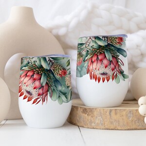 Könnte beinhalten: Zwei weiße, stielose Weingläser mit einem Blumendesign mit roten und grünen Protea-Blüten. Die Gläser stehen auf einer Holzoberfläche, mit dekorativen Vasen und einer Perlenkette im Hintergrund.