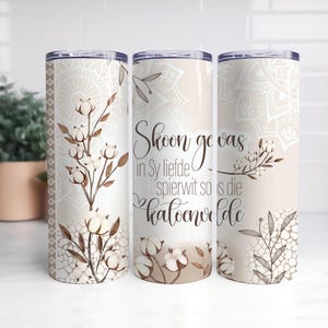 Puede incluir: Tres vasos de acero inoxidable con tapas. Cada vaso presenta un fondo beige con un diseño floral y mandala. El vaso central tiene el texto "Skoon ge vas in Sy liefde spierwit so is die katoenvelde."