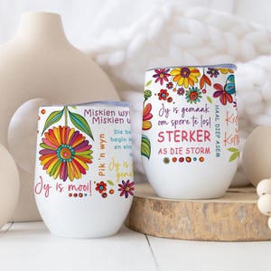 May include: Two white stainless steel tumblers with colourful floral designs and inspirational quotes in Afrikaans. The tumblers are sitting on a wooden surface with a white vase and a string of wooden beads.