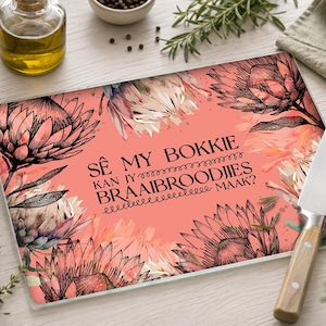 May include: A rectangular glass cutting board with a coral pink background featuring black line art illustrations of protea flowers. The text SÊ MY BOKKIE KAN JY BRAAIBROODJIES MAAK? is printed in black in the center. A silver knife with a wooden handle rests on top.