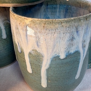 May include: A stack of handmade ceramic mugs. The mugs are teal with a white glaze that drips down the sides. Each mug has a handle and a rounded shape. The mugs are stacked on top of each other.