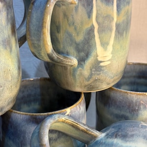 May include: Several handmade ceramic mugs with a blue and tan glaze. The mugs have large, curved handles and a glossy finish. The color palette includes shades of blue, tan, and cream. The mugs are likely for drinking tea or coffee.