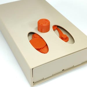 Op de afbeelding: Een beige doos met een lade, met oranje mechanische componenten. De bovenkant heeft ovale uitsparingen die oranje tandwielen, een cilindrisch stuk en een gebogen hendel onthullen. Het ontwerp suggereert een puzzel of een mechanisch speelgoed.