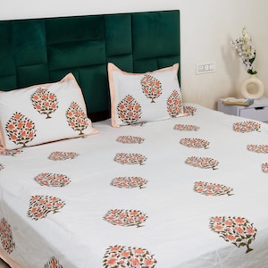 May include: White bed linen set featuring a repeating floral design in orange and green. The set includes two pillowcases and a fitted sheet. The pillowcases have an orange striped border. The bed is against a dark green headboard.