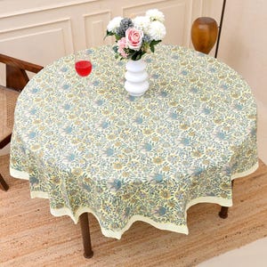 Könnte beinhalten: Eine runde Tischdecke mit einem Blumenmuster in Blau-, Gelb- und Grüntönen. Die Tischdecke liegt auf einem runden Tisch mit dunkelbraunen Beinen. Eine Vase mit Blumen und ein Glas mit roter Flüssigkeit stehen auf dem Tisch.