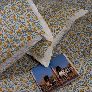 Peut inclure: Ensemble de literie avec un motif floral de fleurs jaunes et de feuilles bleu-vert sur un fond crème. Les taies d'oreiller ont une bordure rayée. Un livre ouvert avec une photographie d'un chariot est sur le lit.