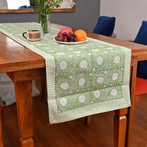 May include: A green and white floral table runner with a striped border rests on a wooden table. A cup, a plate of fruit, and a vase of flowers are on the runner. The runner's design features white flowers on a green background.