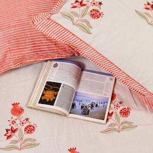 May include: White and red floral patterned bedding set with a book open in the middle. The bedding features red pomegranate and floral designs. The pillowcase has red and white stripes.