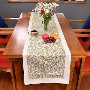 Può includere: Un tavolo da pranzo in legno con un runner a motivi floreali. Il runner presenta un bordo bianco e un disegno di fiori gialli e foglie verdi. Un vaso di fiori, una tazza e un piatto di frutta sono sul tavolo.