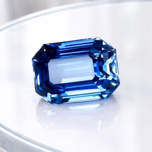 Op de afbeelding: Een gefacetteerde, rechthoekig geslepen edelsteen met een diepblauwe kleur. De facetten van de steen reflecteren het licht, waardoor een schitterend effect ontstaat. De edelsteen staat tegen een witte achtergrond. Geschikt voor sieraden.