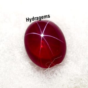 May include: A polished, oval-shaped, deep red gemstone with a six-rayed star pattern. The gem has a high luster and is set against a white, textured background. The word "Hydragems" is written above the gem.