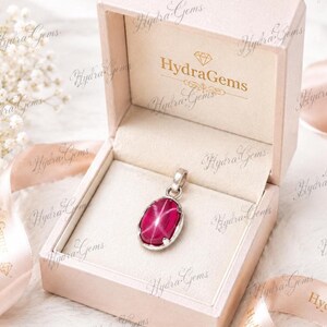 Può includere: Un ciondolo ovale in rubino stellato rosa intenso incastonato in una montatura in metallo color argento, esposto all'interno di un portagioie aperto di colore chiaro con il testo HydraGems stampato in oro sul coperchio e un nastro abbinato.