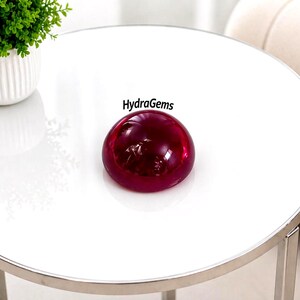 May include: A large, round, polished, deep red gemstone sits on a white table. The gemstone has a smooth, reflective surface. The table is round with a silver metal trim. The text "HydraGems" is visible.