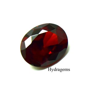 Peut inclure: Une pierre précieuse rouge foncé de forme ovale avec une surface à facettes qui reflète la lumière. La pierre présente une finition polie et une teinte cramoisie profonde. Le texte Hydragems est imprimé en noir dans le coin inférieur droit.