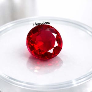 May include: A vibrant red, round-cut gemstone, likely a ruby, is displayed on a white surface. The gem's facets catch the light, highlighting its brilliance. The text "HydraGems" is present.