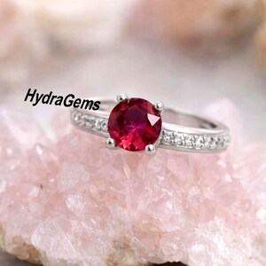 Puede incluir: Un anillo de plata con una gran gema redonda de color rojo intenso como pieza central. La banda está adornada con gemas más pequeñas y transparentes. El anillo está sobre un fondo rosa claro y texturizado. El texto "HydraGems" es visible.