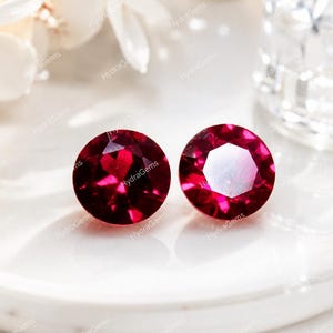 Könnte beinhalten: Zwei runde, facettierte, tiefrote Edelsteine im Brillantschliff. Die Steine zeigen einen leuchtend karmesinroten Farbton und reflektierende Oberflächen mit zahlreichen dreieckigen und sechseckigen Facetten, die das Licht einfangen.
