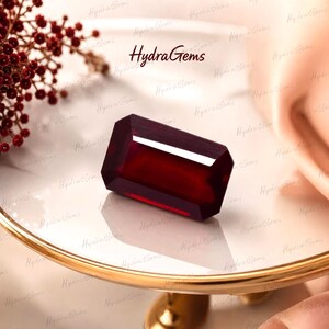 Op de afbeelding: Een rechthoekige, gefacetteerde, dieprode edelsteen. De edelsteen wordt weergegeven op een goudomrand, wit oppervlak, met de tekst "Hydra Gems" zichtbaar. Decoratieve rode bessen zijn op de achtergrond.