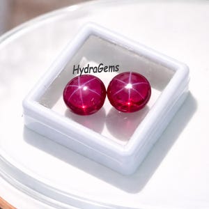 Könnte beinhalten: Zwei runde, tiefrote Edelsteine mit Sterneneffekt, präsentiert in einer weißen quadratischen Box. Die Edelsteine haben eine polierte, reflektierende Oberfläche. Der Text "HydraGems" ist auf der Box zu sehen. Diese Edelsteine eignen sich für die Schmuckherstellung.