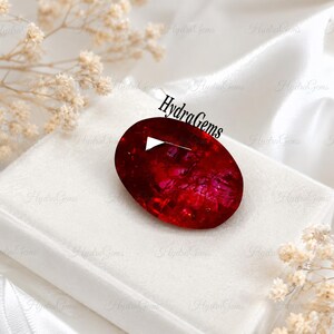 May include: An oval-shaped, deep red gemstone with a faceted surface and internal light reflections. The stone features a prominent white triangular highlight on its upper left side and the text HydraGems printed in black above it.