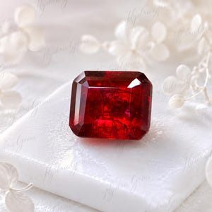 Può includere: Una gemma rosso intenso e vibrante con taglio a smeraldo rettangolare, caratterizzata da bordi sfaccettati netti e un interno cristallino traslucido con inclusioni visibili, appoggiata su una superficie bianca, quadrata e liscia.