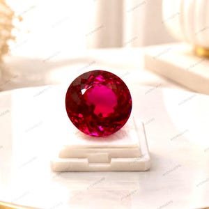 May include: A round, faceted, vibrant red gemstone with a brilliant cut, displaying deep crimson hues and light-reflecting surfaces. The stone is securely positioned on a small, square, white display stand, highlighting its intense color and clarity.